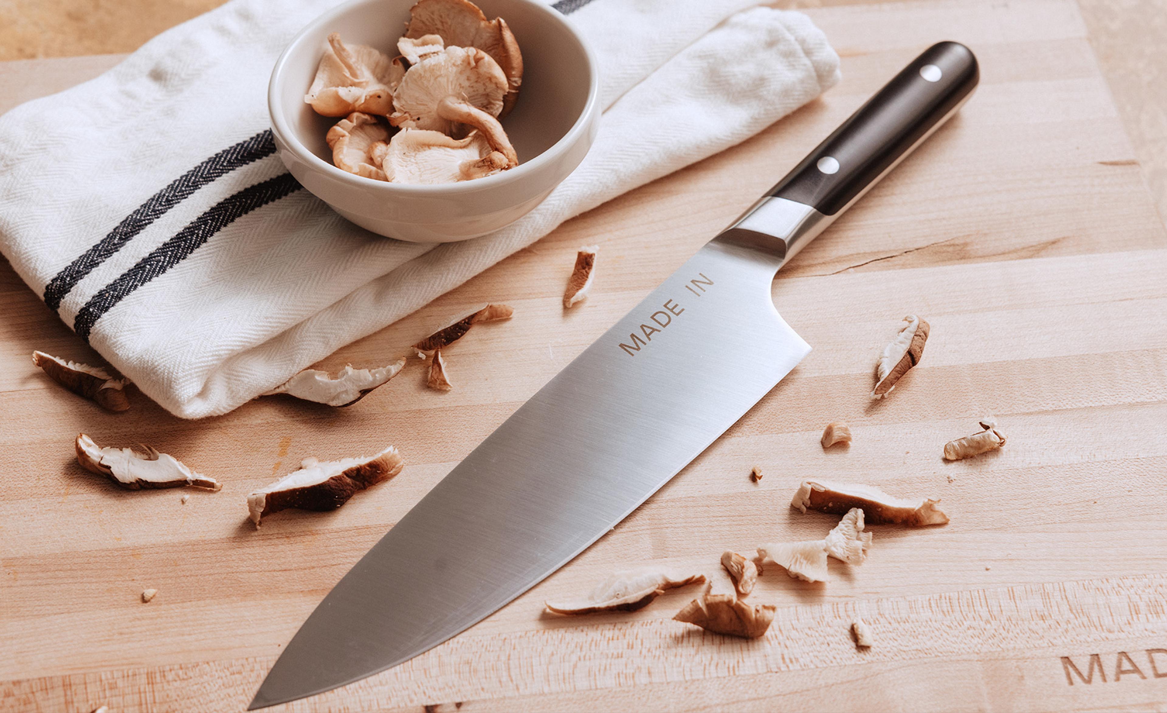 A stainless steel chef's knife rests on a wooden cutting board next to a bowl of sliced mushrooms and scattered mushroom pieces.