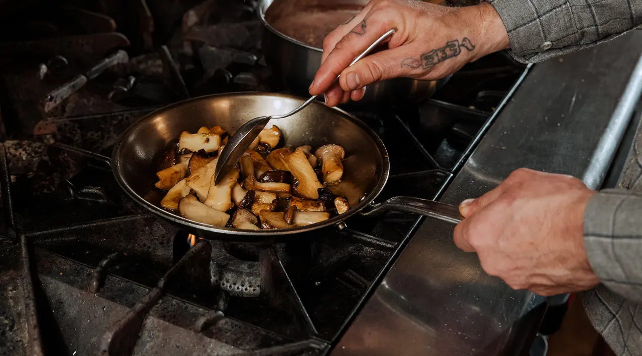 A person with tattooed arms is sautéing food in a pan over a stove.