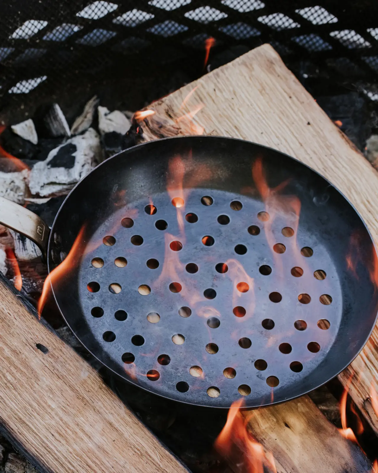 A perforated pan sits atop a wood fire, with flames licking through the holes.