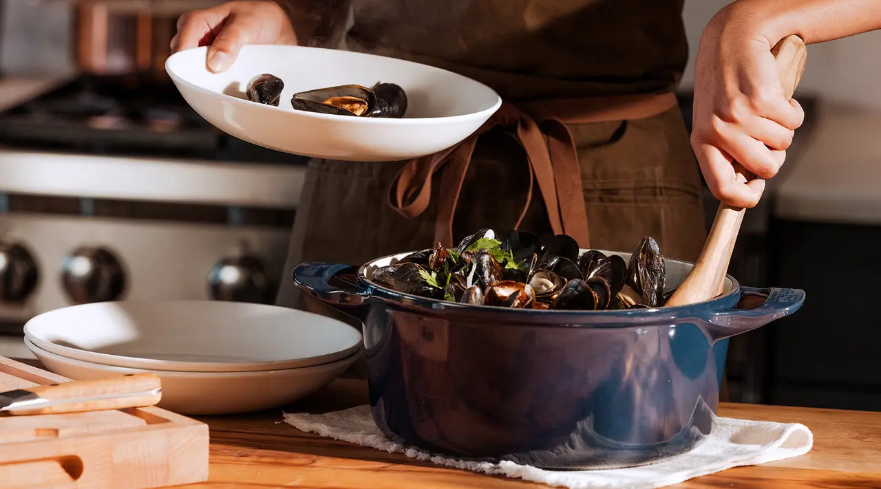 A person cooks mussels in a blue pot on a kitchen stove, while holding a bowl.