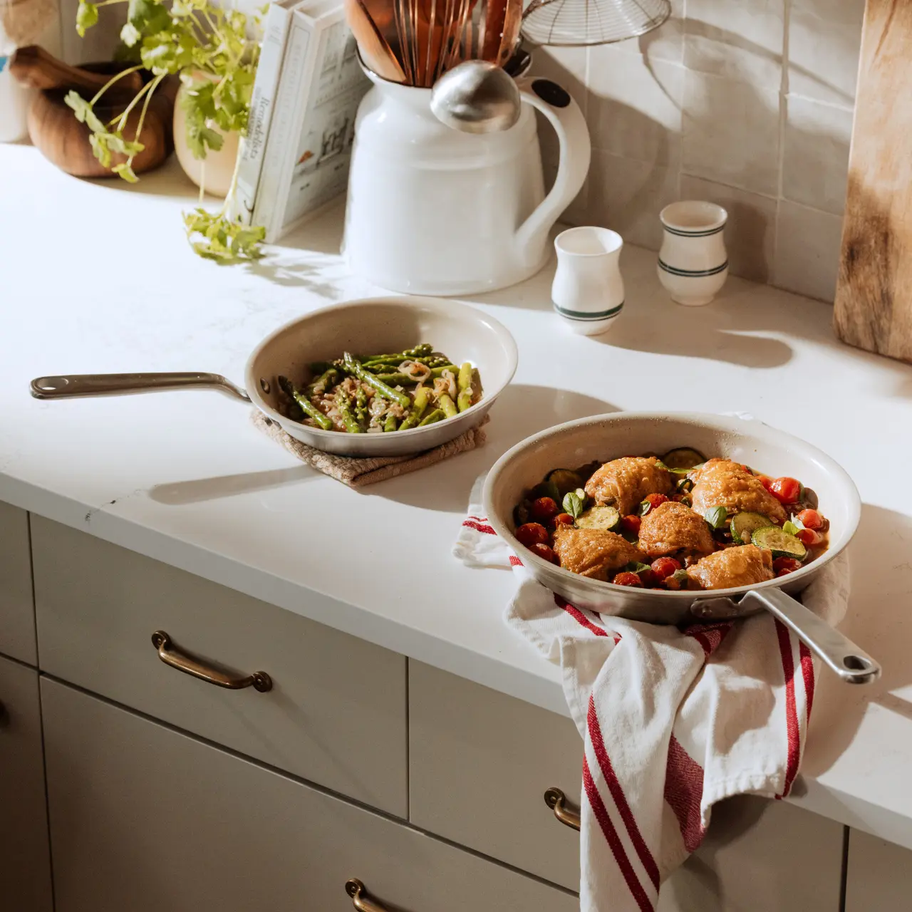 Two serving dishes sit on a countertop: one with roasted vegetables and another with asparagus, accompanied by kitchen utensils and a small plant.
