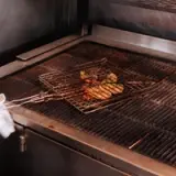 Grilled vegetables and meat are cooking on a wire rack over a flame in a commercial kitchen setting.