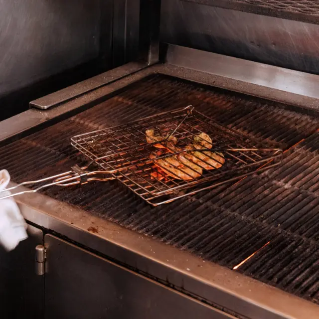 Grilled vegetables and meat are cooking on a wire rack over a flame in a commercial kitchen setting.