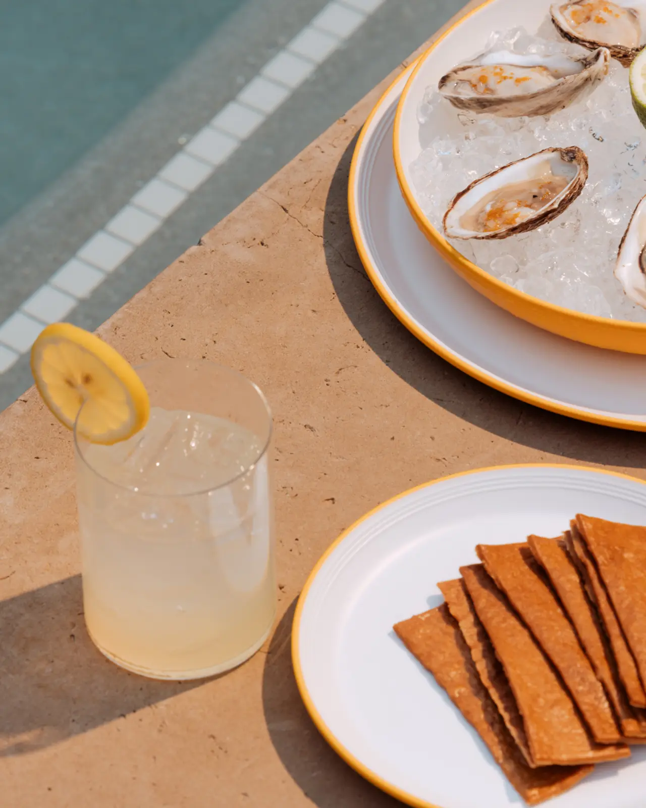 A refreshing beverage with a lemon slice accompanies a platter of oysters on ice and crisp crackers by a poolside.