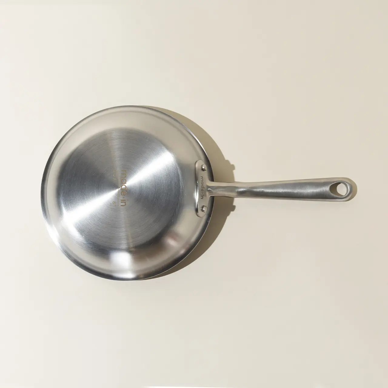 A stainless steel frying pan with a long handle is mounted on a pale background, casting a soft shadow.