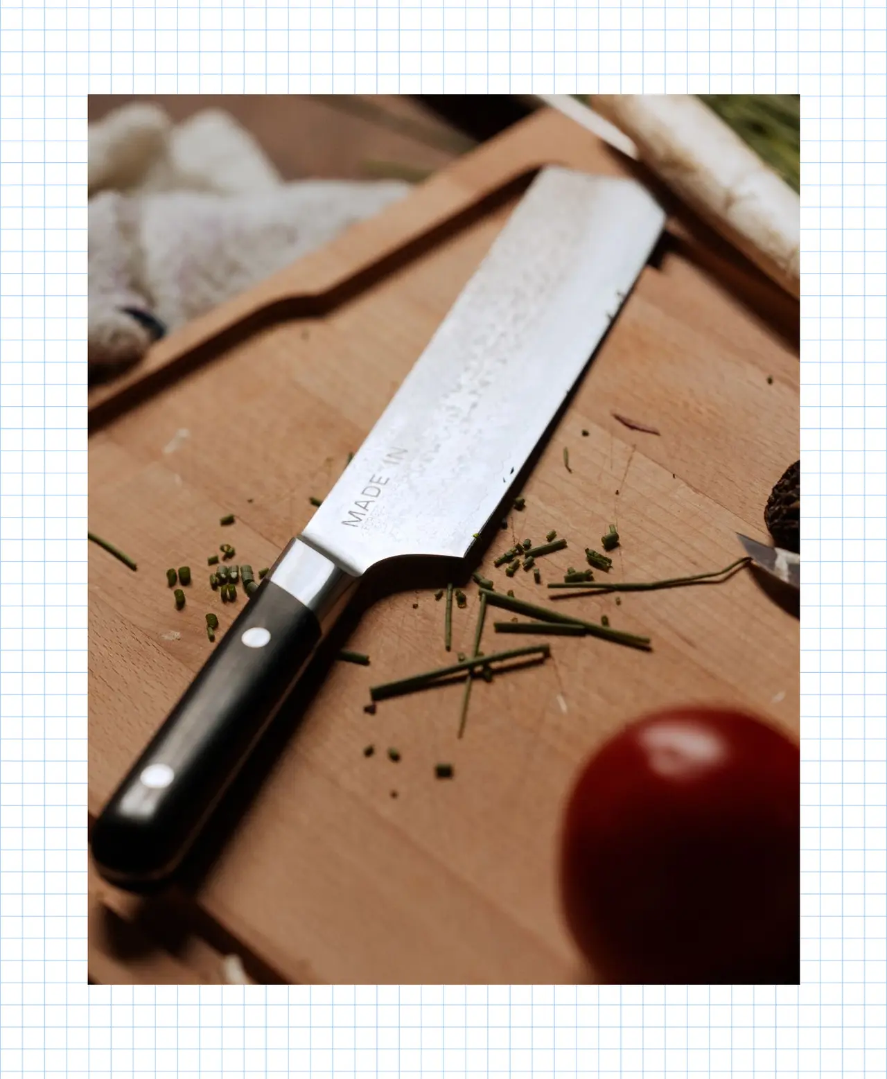 A chef's knife lies on a wooden cutting board surrounded by chopped herbs and a whole tomato, suggesting food preparation is underway.