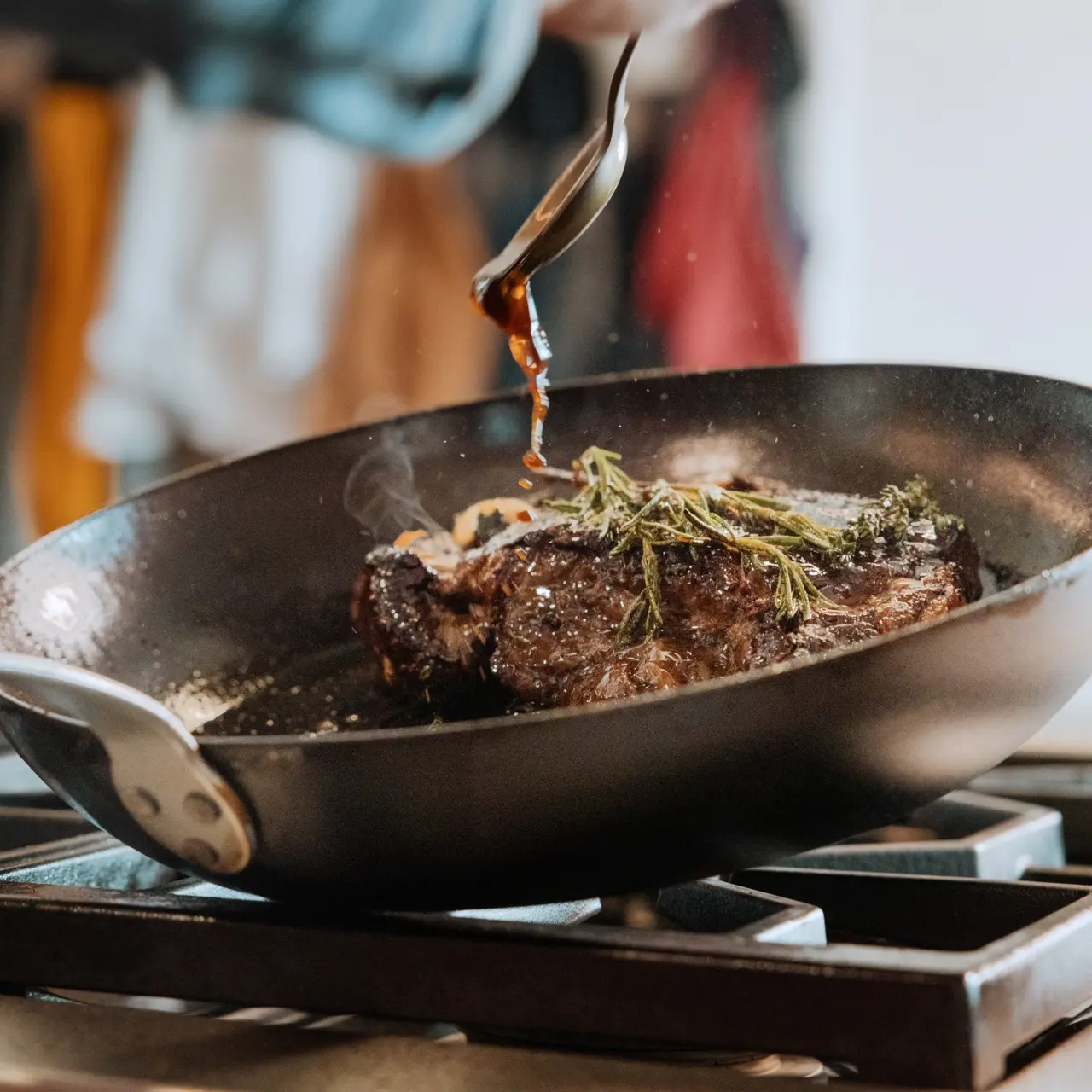 A close-up view of a sizzling steak being cooked in a frying pan, garnished with herbs and dripping with juices.