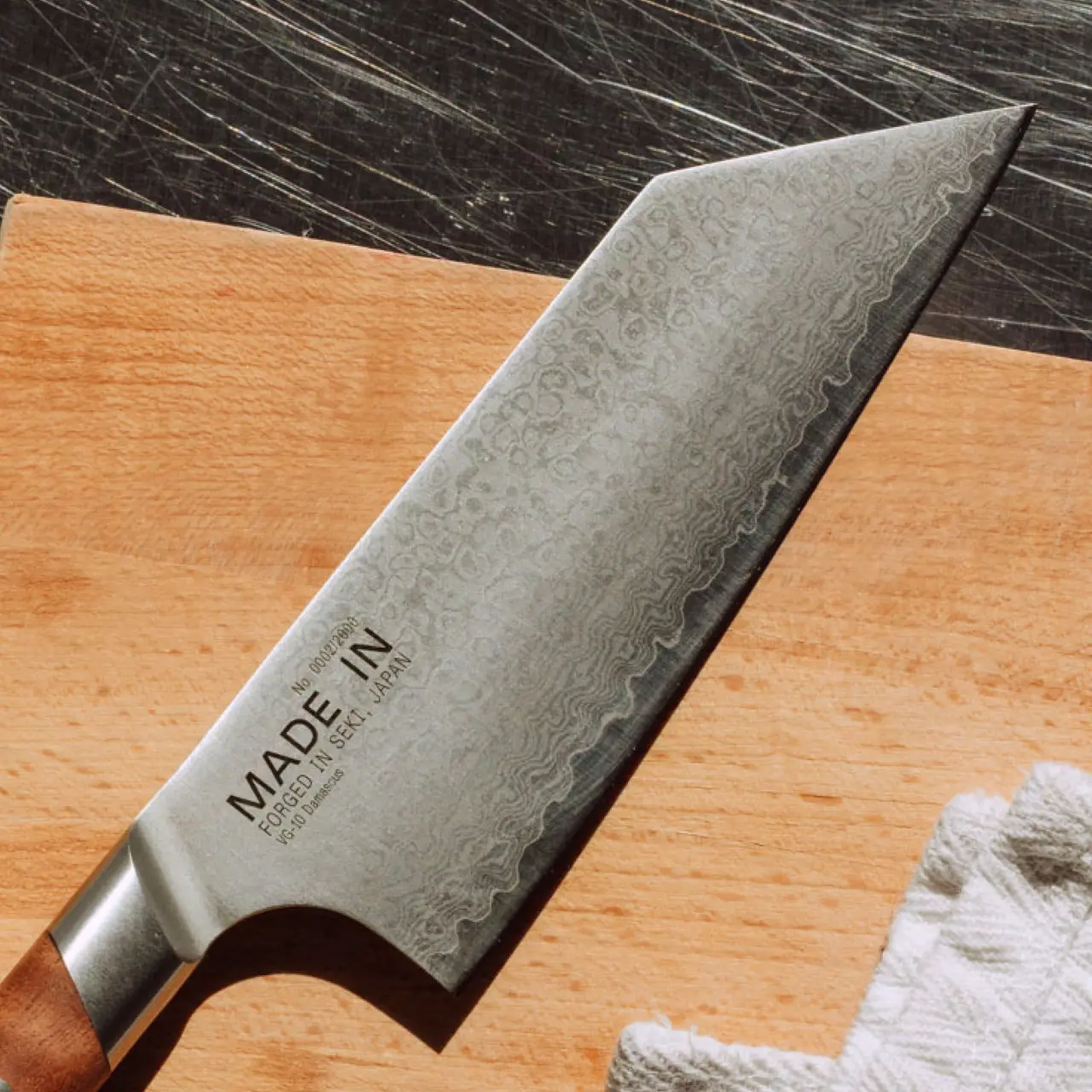 A well-crafted chef's knife with a distinctive patterned blade rests on a wooden cutting board alongside a white kitchen towel.