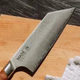 A well-crafted chef's knife with a distinctive patterned blade rests on a wooden cutting board alongside a white kitchen towel.