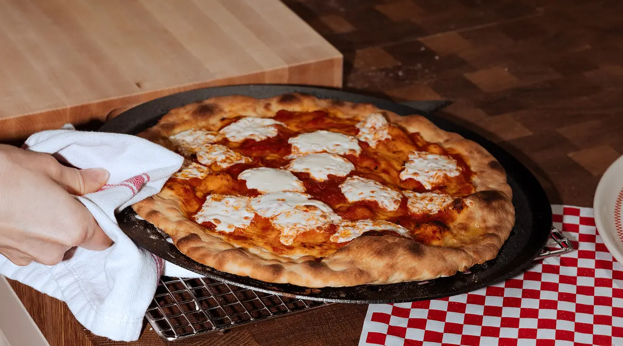 A freshly baked pizza with bubbly cheese is being served on a wooden table with a classic red and white checkered tablecloth.