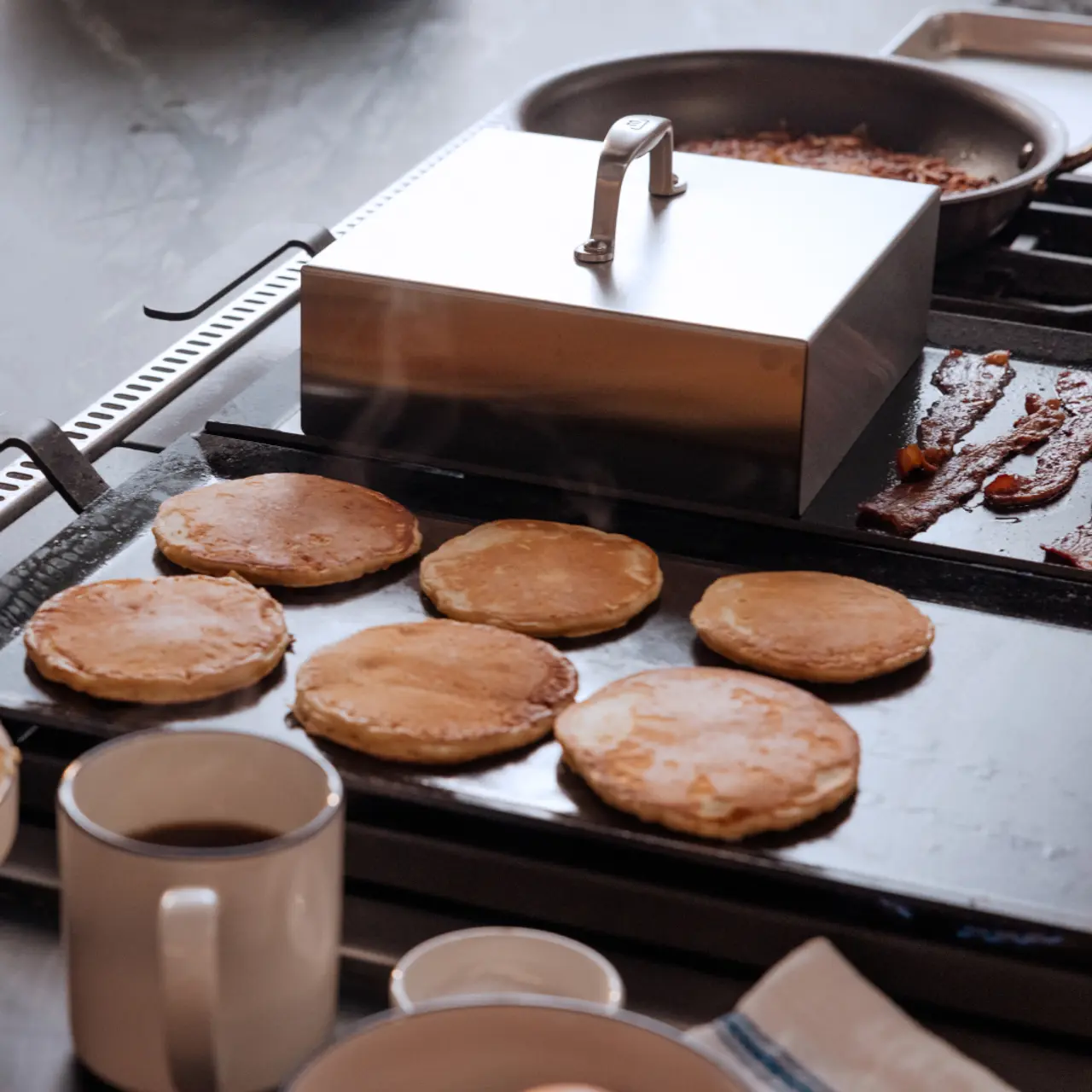 A griddle cooks pancakes beside a skillet with bacon and a mug, conveying a breakfast preparation scene.