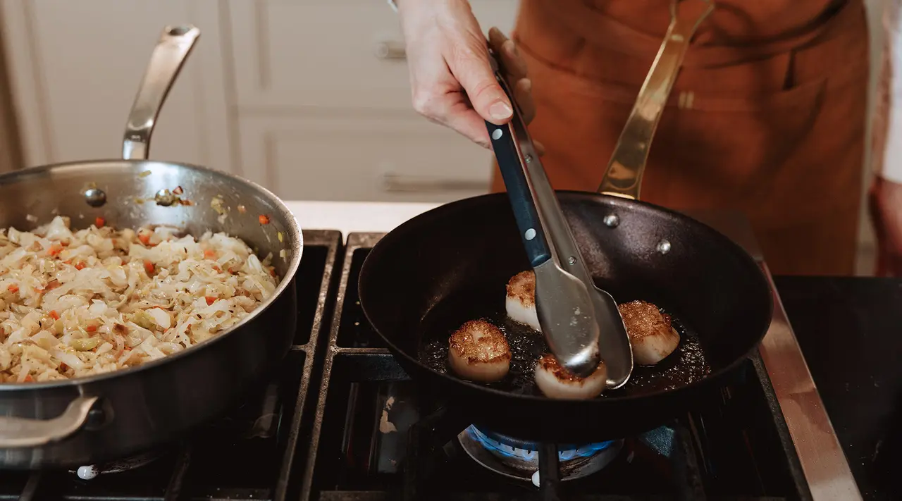 A person in an apron uses tongs to sauté scallops in a frying pan while a pot of vegetable rice cooks on the stove next to them.