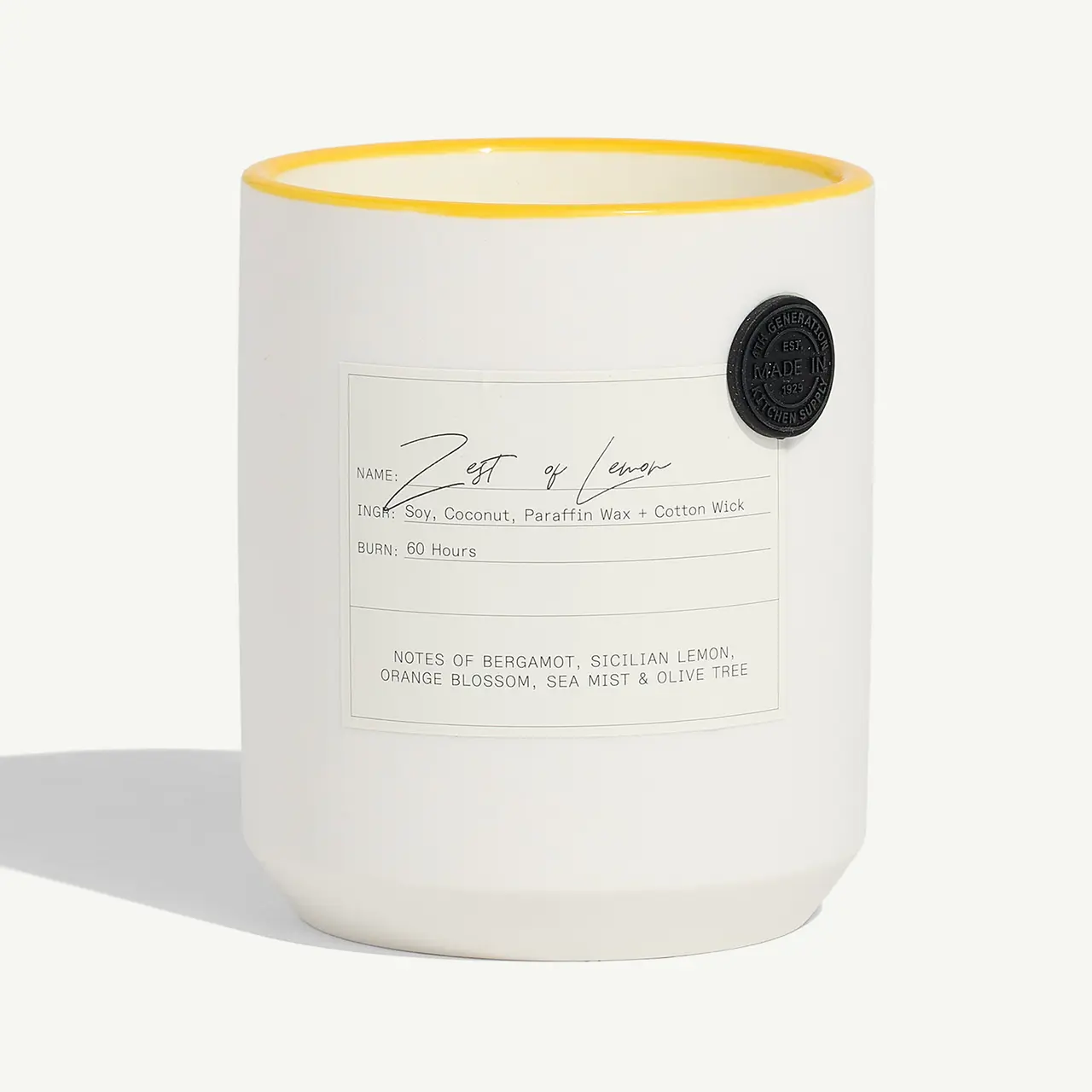 A white candle with a yellow rim and a description label detailing its scent components and burn time, alongside a black circular emblem.