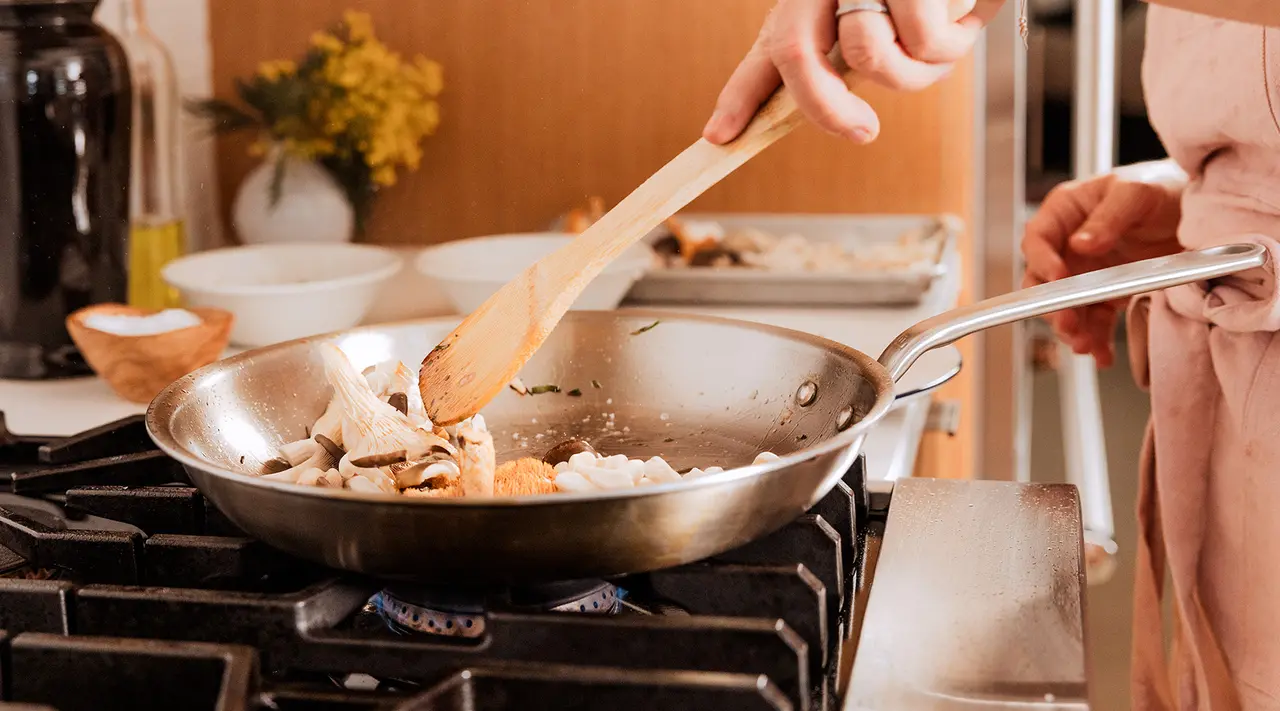 A person is cooking in a pan on a stove using a wooden spatula, with kitchen items and ingredients in the background.