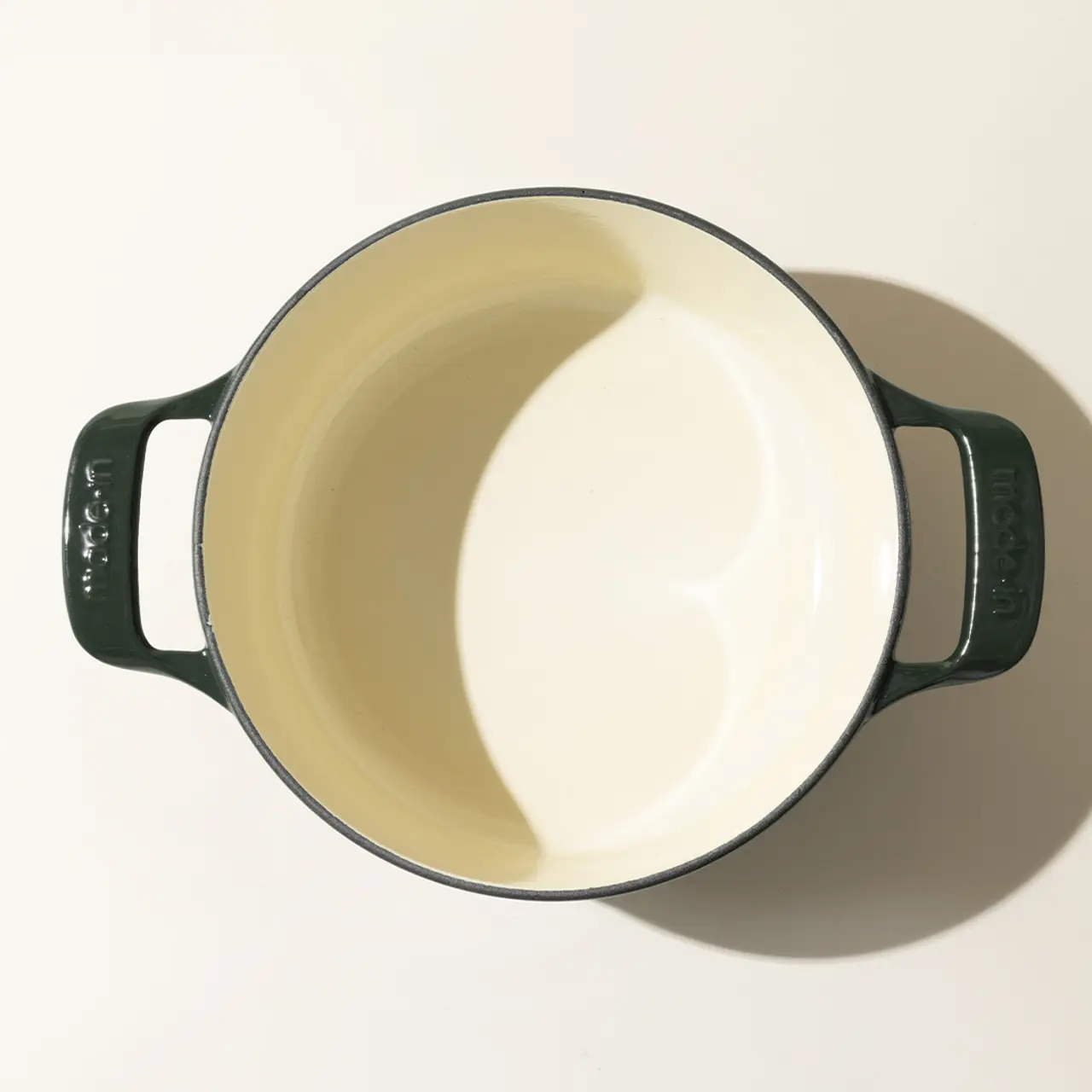 An empty, cream-colored enamel pot with two dark green handles viewed from directly above, casting a soft shadow on a light surface.