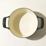 An empty, cream-colored enamel pot with two dark green handles viewed from directly above, casting a soft shadow on a light surface.