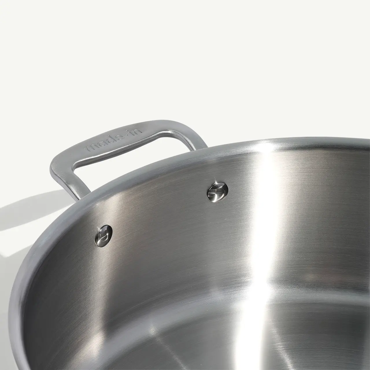 A close-up view of a stainless steel pot with its riveted handle visible against a neutral background.