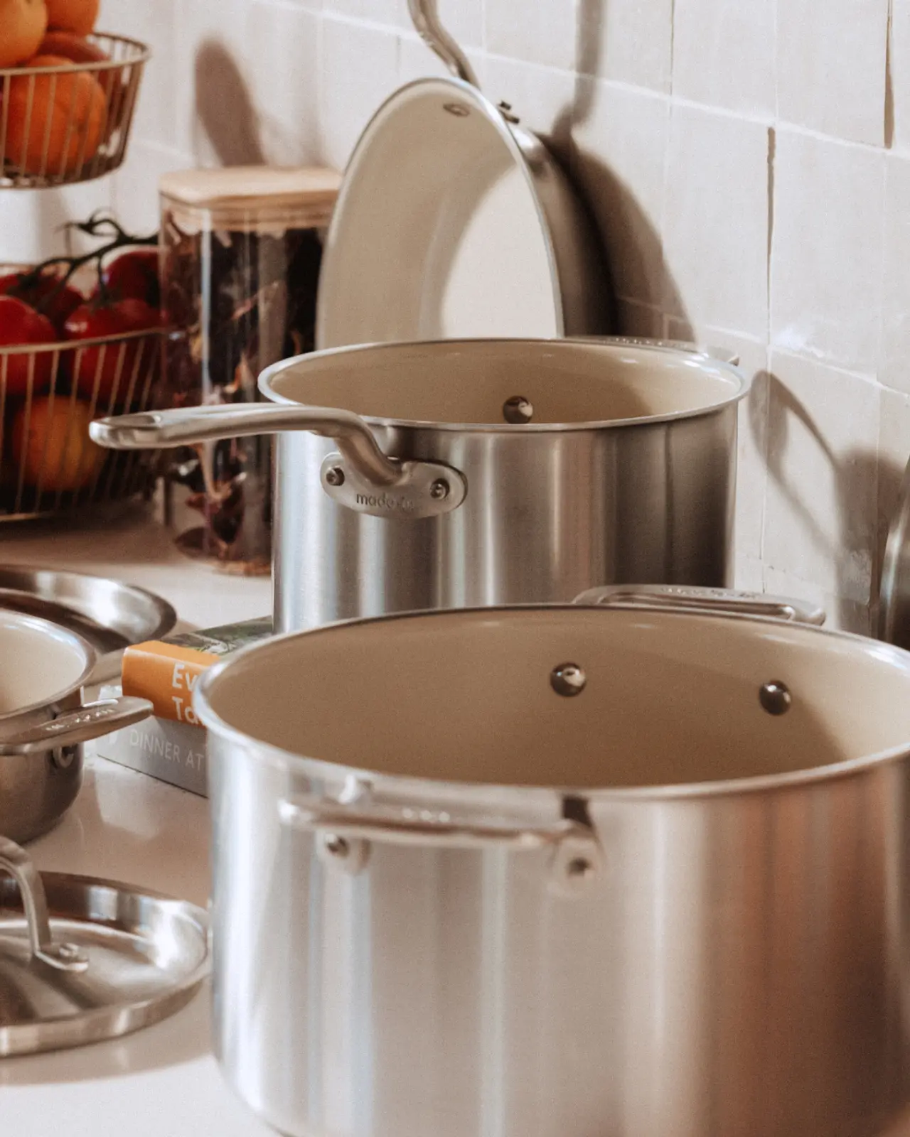 Two stainless steel cooking pots are placed on a kitchen countertop next to a lid and a bowl, with a basket of fruits in the background.