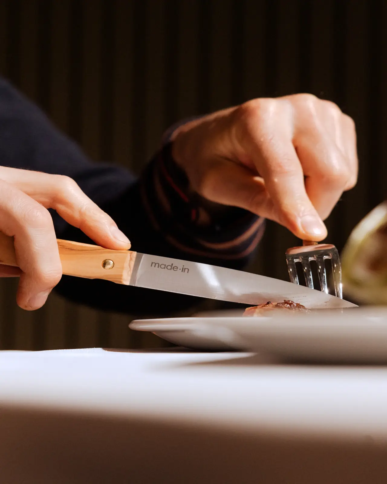 Close-up of a person's hands using a fork and knife to cut meat on a plate at a dining table.