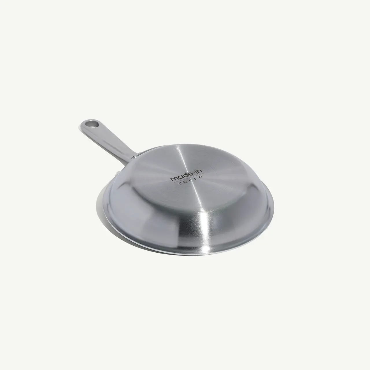 A stainless steel frying pan is positioned upside-down on a light background, showcasing its bottom and handle.