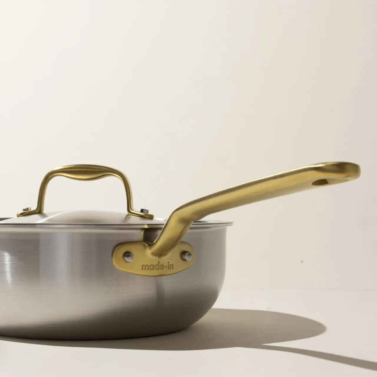 A stainless steel saucepan with a gold-colored handle and lid handle set against a neutral background.