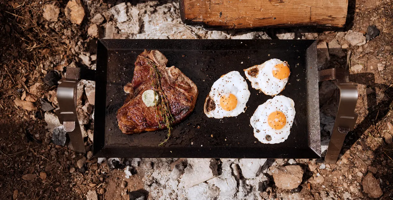 A top-down view of a cooking surface over embers with a steak and three fried eggs.