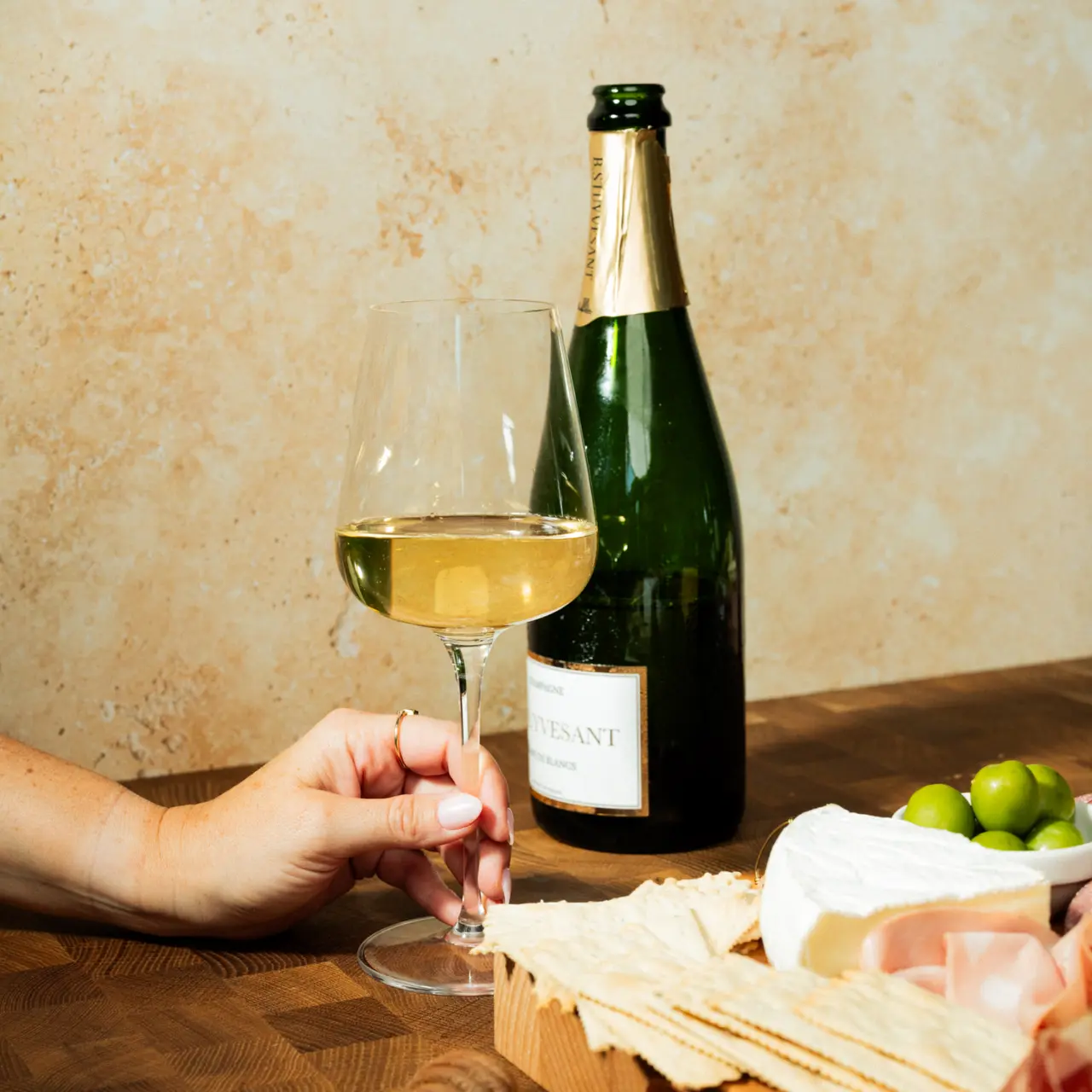 A hand holds a stemmed glass of white wine near a charcuterie board with cheese and meats, with a bottle in the background.