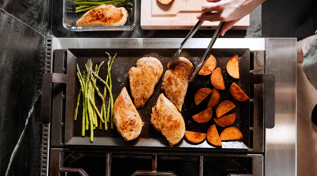 A person is cooking chicken breasts, sweet potatoes, and asparagus on a stove-top grill.