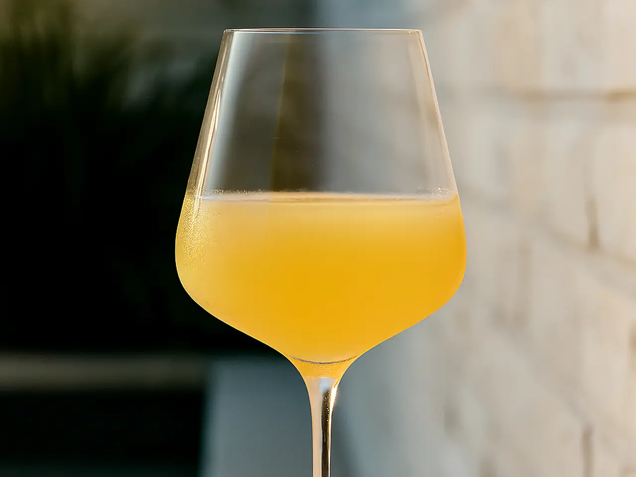 A crystal-clear wine glass filled with a vibrant yellow-orange beverage is set against a softly lit, textured wall.