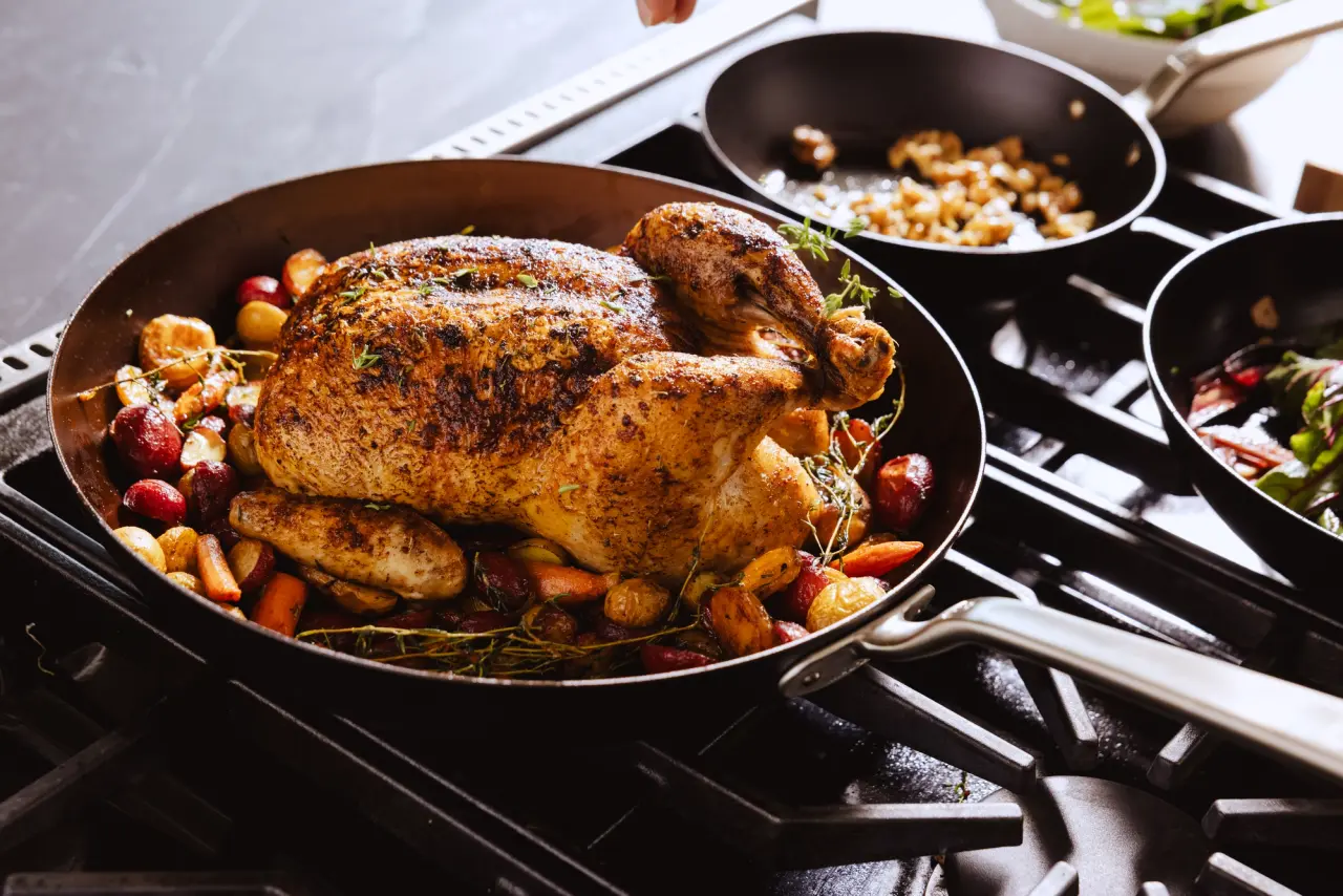 A seasoned roast chicken is showcased in a skillet, surrounded by colorful vegetables and herbs, with additional pans of food in the background.
