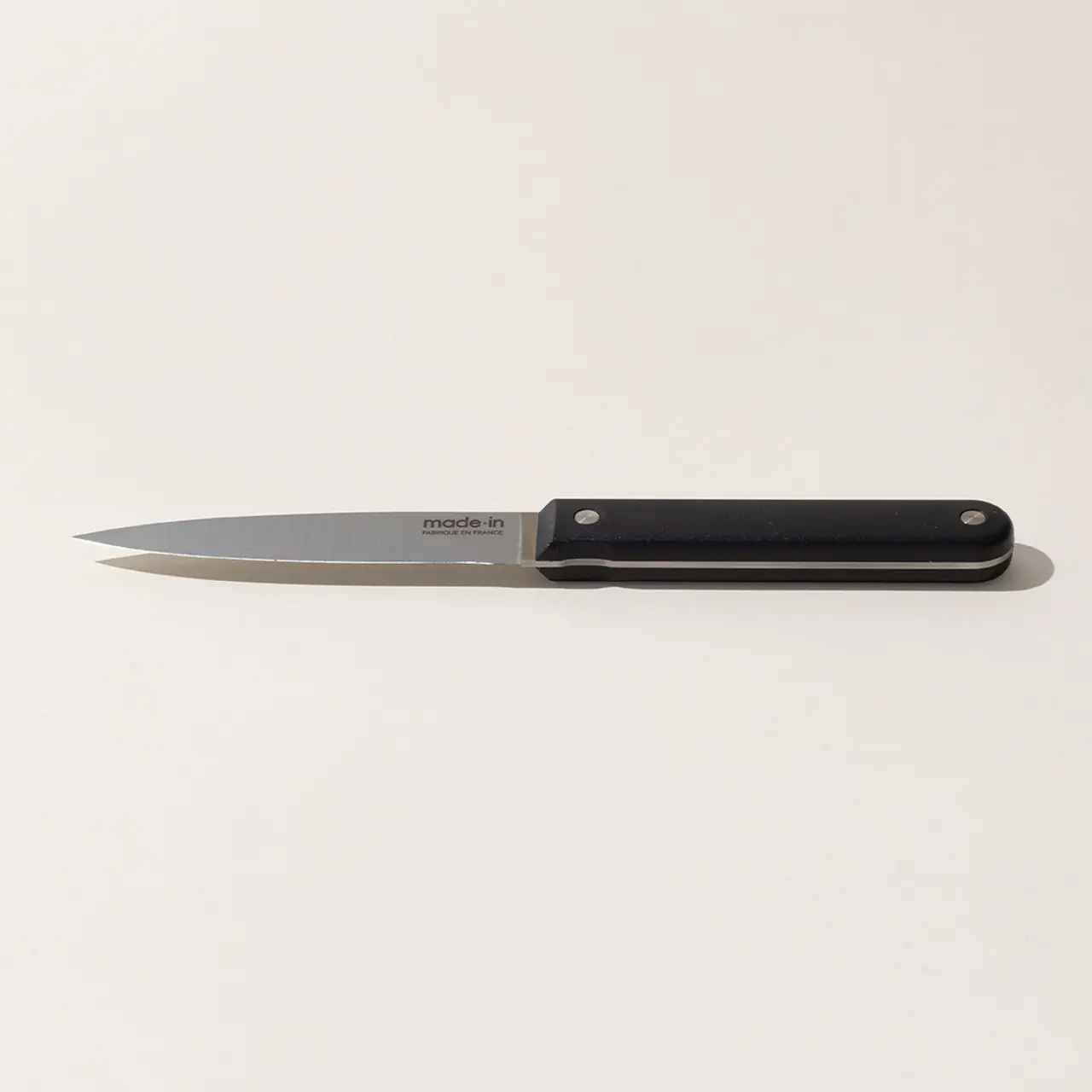 A kitchen knife with a black handle lies on a plain, light-colored surface.
