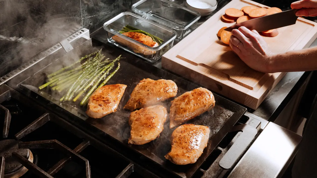A person is cooking chicken breasts on a griddle while slicing vegetables on a cutting board nearby, with steam rising from the sizzling food.