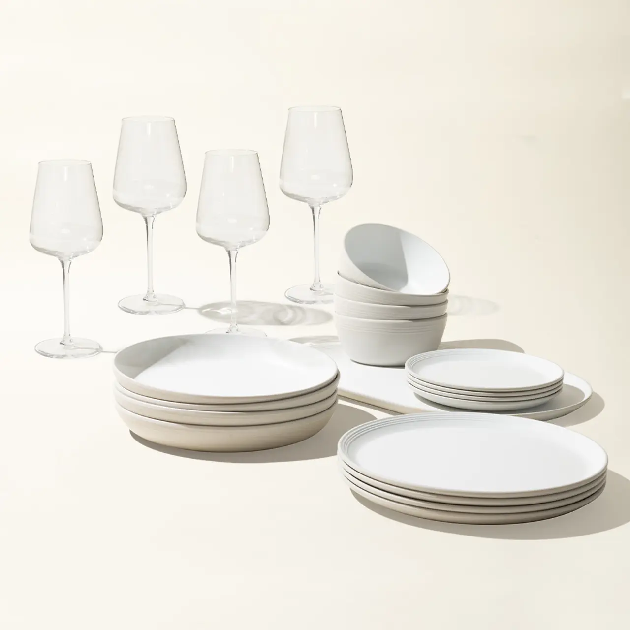 A collection of elegant white dishes and clear wine glasses are neatly arranged on a light background.