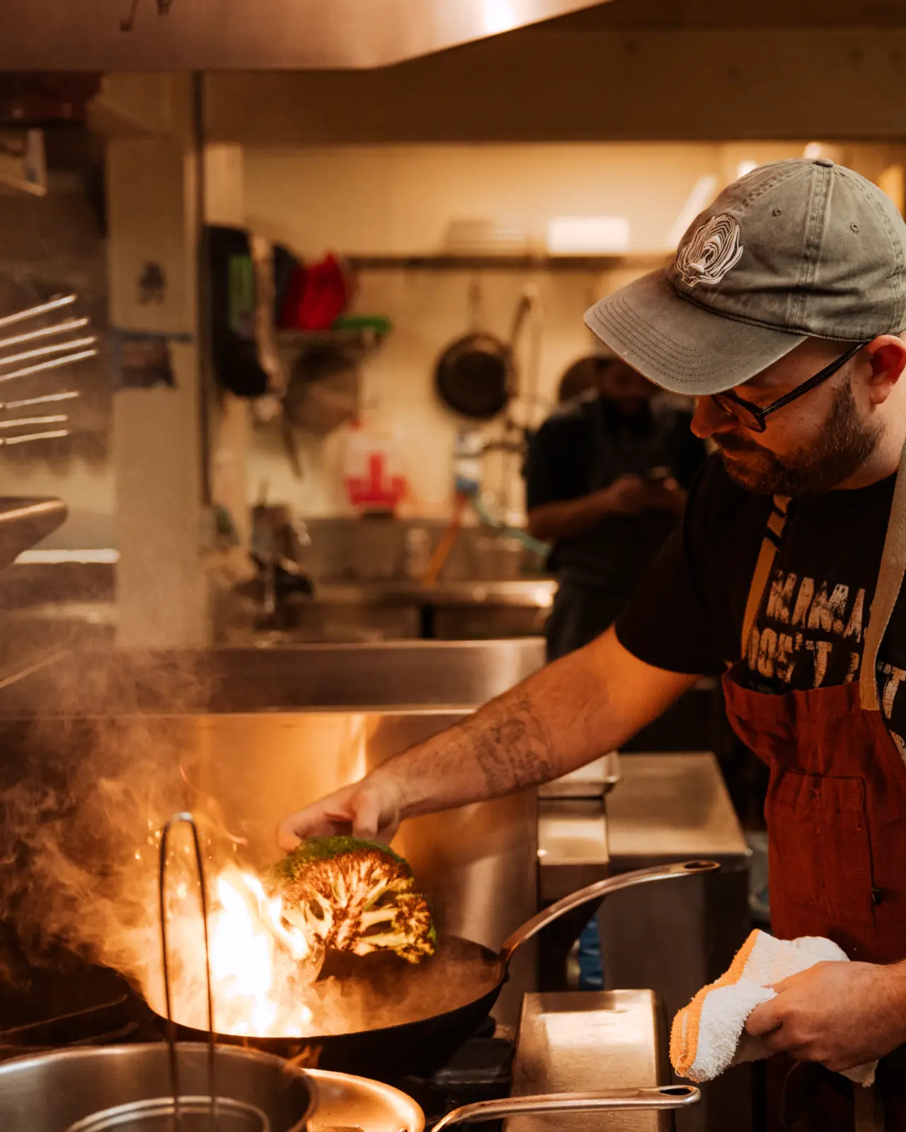 A chef wearing a baseball cap and an apron attentively cooks with a flaming pan in a bustling kitchen environment.