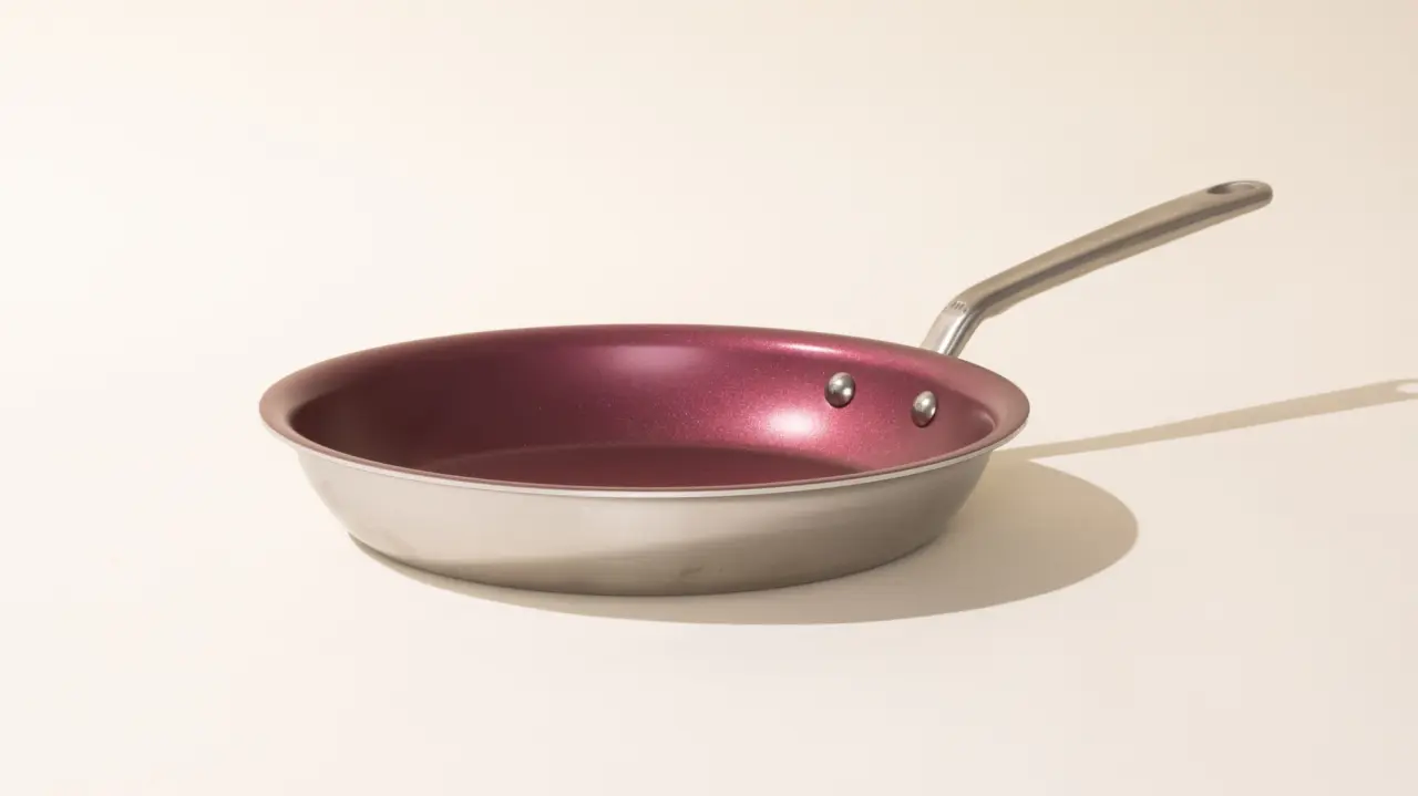 A stainless steel frying pan with a maroon interior resting on a neutral surface casting a soft shadow.