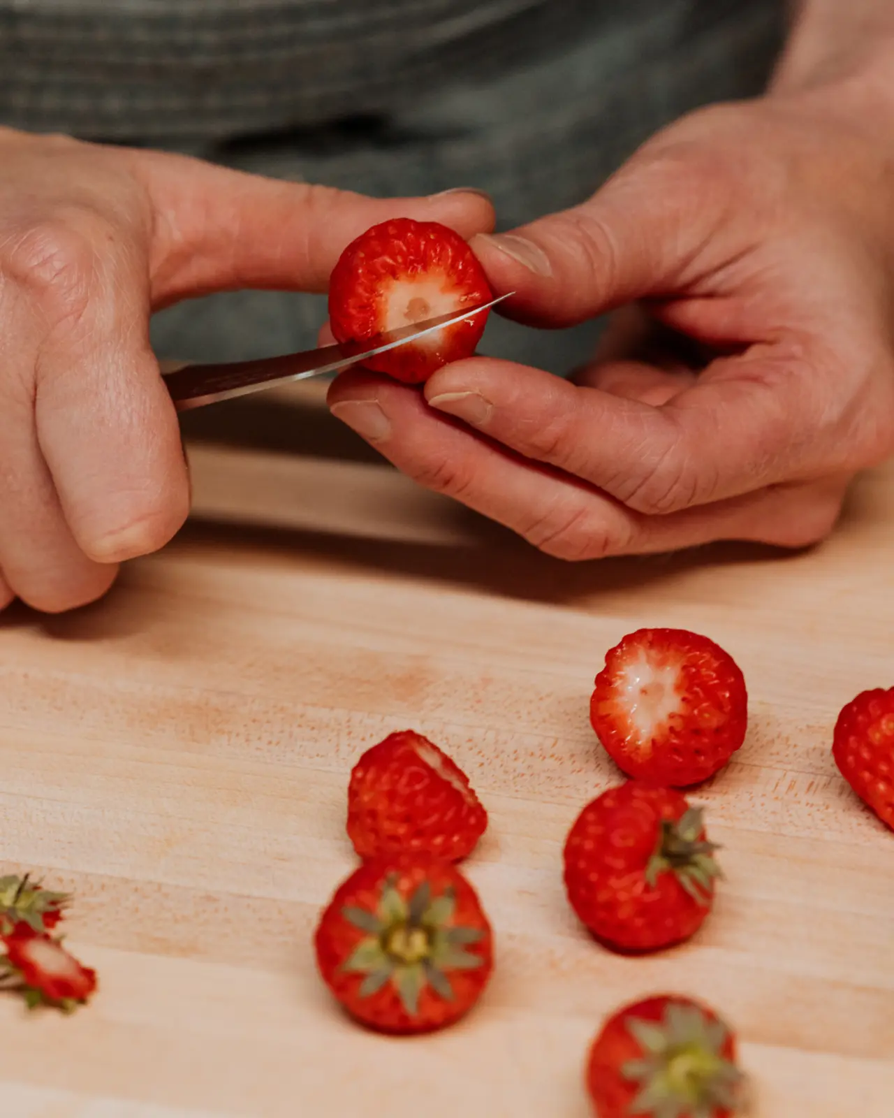 A person is slicing a bright red strawberry on a wooden board with several other strawberries around it.