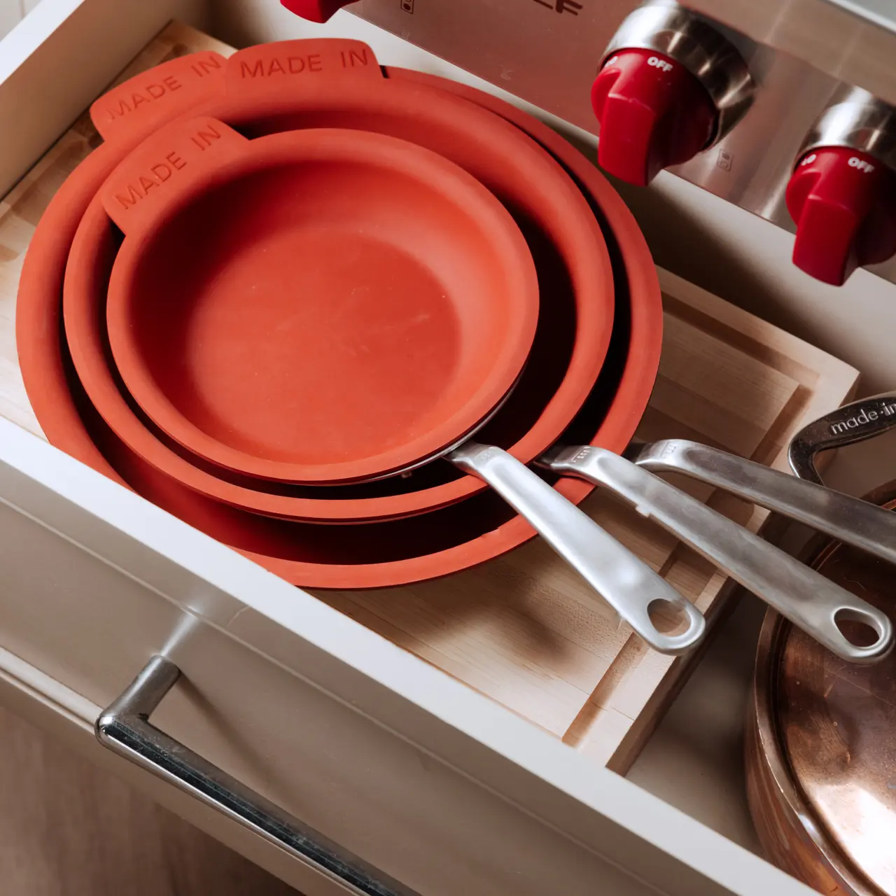 A set of red-orange pans is neatly stacked in an organized kitchen drawer beside a stainless steel pot with red knobs.