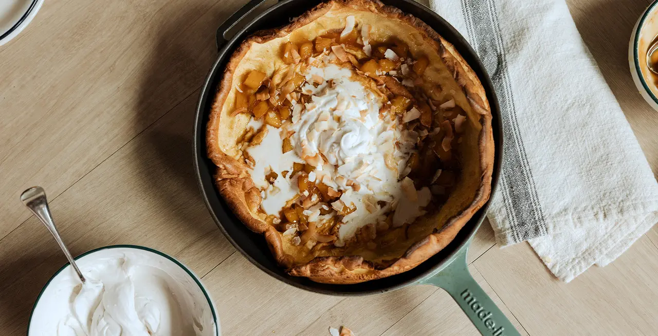 A freshly baked Dutch baby pancake topped with whipped cream and nuts in a skillet, next to a bowl of whipped cream.