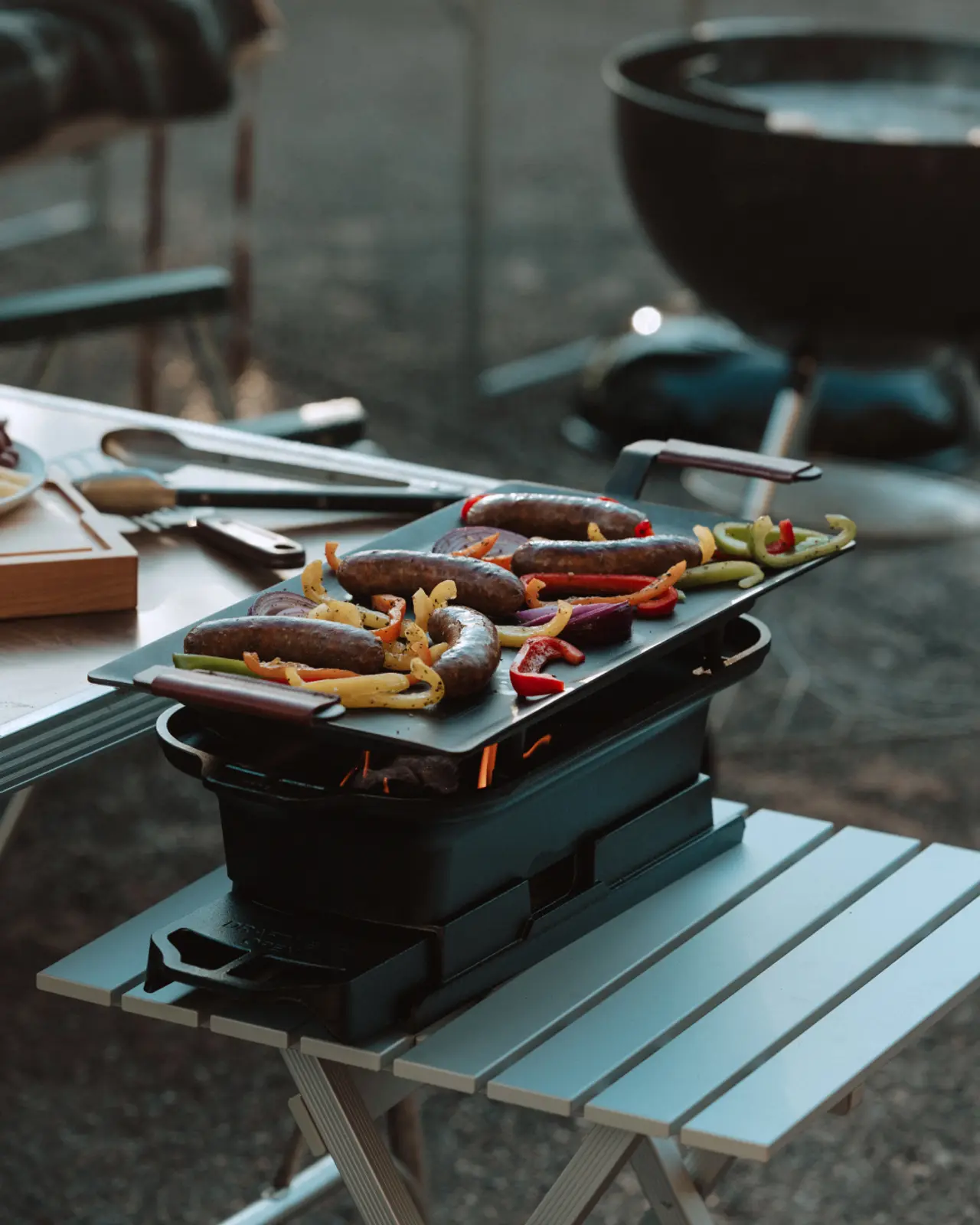 A portable grill with various sausages and sliced vegetables cooking on top is set upon a compact outdoor table.