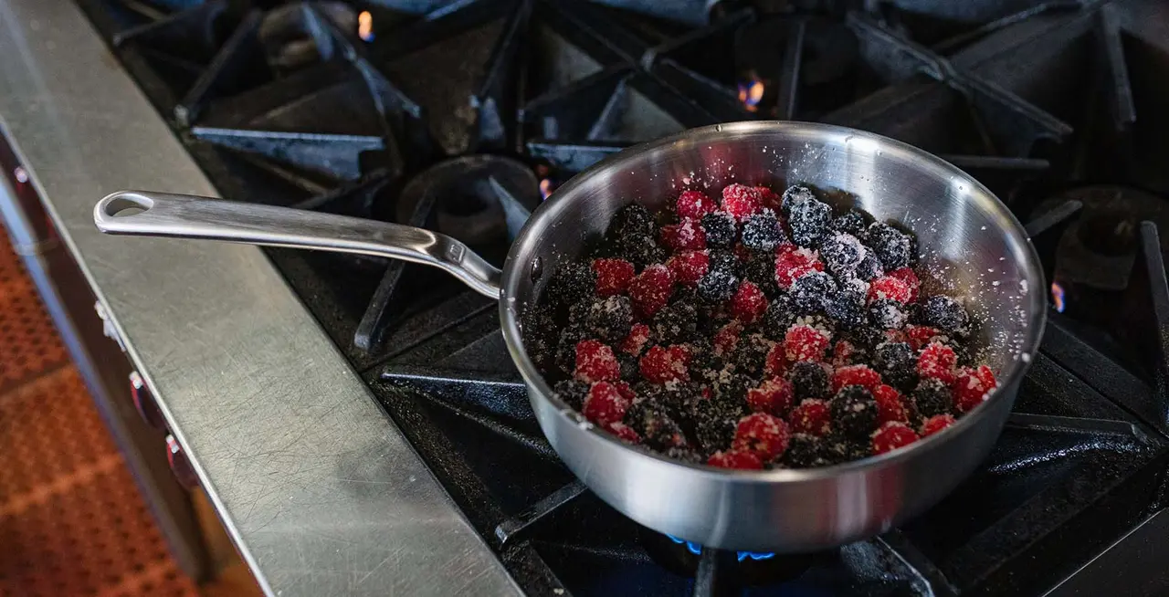 A stainless steel skillet on a gas stove contains a mix of cooking blackberries and raspberries.