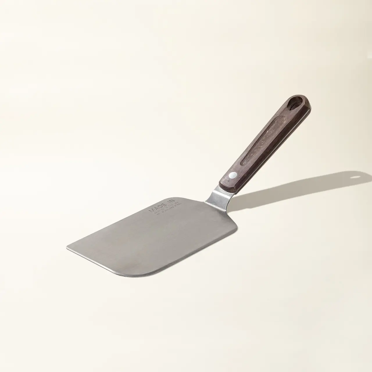 A stainless steel cheese knife with a riveted wooden handle is lying isolated on a pale background.