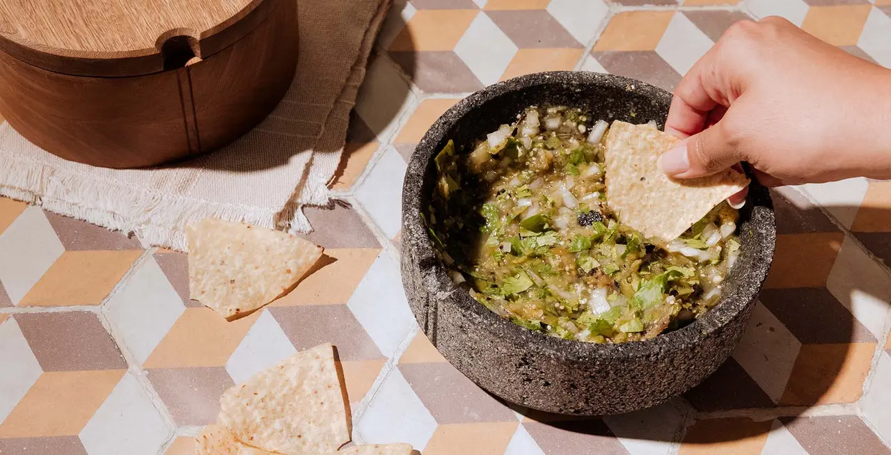 A hand is dipping a tortilla chip into a stone bowl of salsa on a tiled table next to a wooden basket.