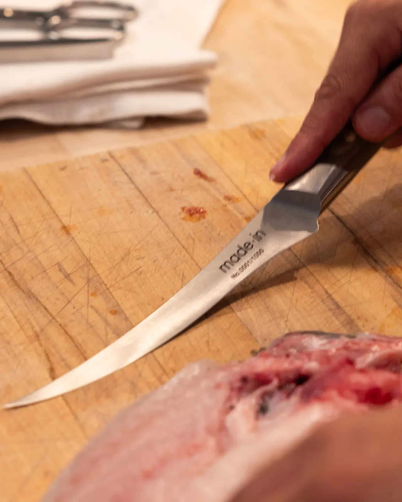 A person's hand is holding a boning knife over a wooden cutting board next to some meat with visible blood spots.