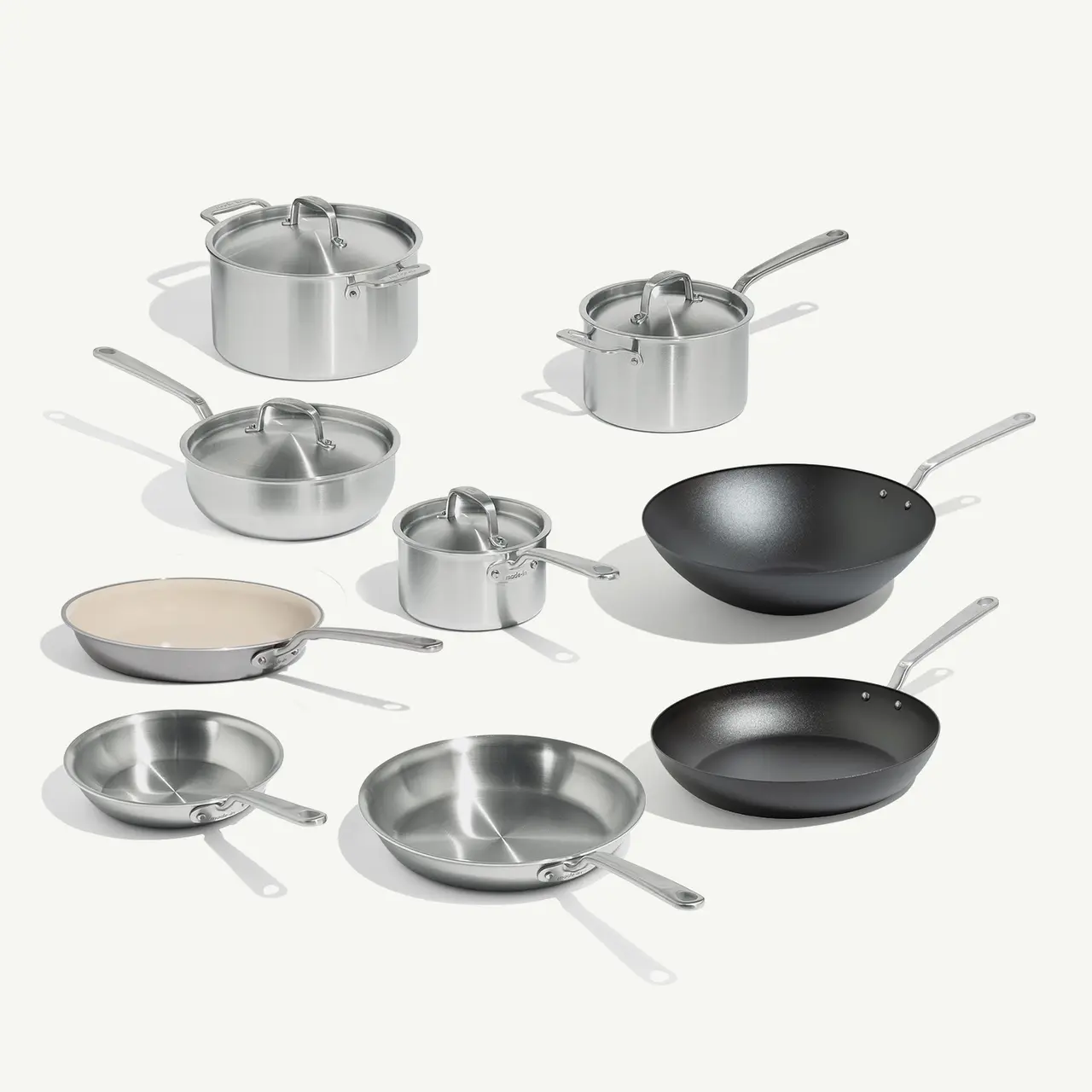 A collection of various cooking pots and pans in stainless steel and one black non-stick wok, arranged aesthetically on a light background.