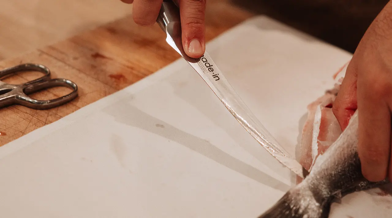 A close-up captures a person's hand filleting a fish with a sharp knife on a cutting board, with a pair of scissors lying nearby.