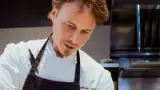 A focused chef wearing a white coat with his name embroidered on it is intently preparing food in a professional kitchen.