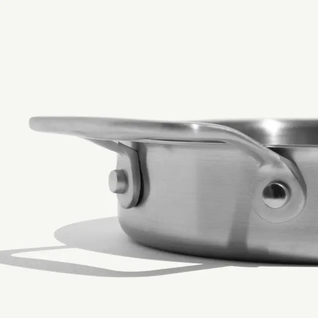 A shiny stainless steel pan with a curved handle is displayed against a light background, casting a shadow.