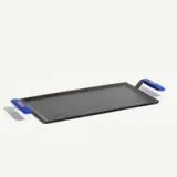 A black griddle with blue silicone handles is positioned on a light background.