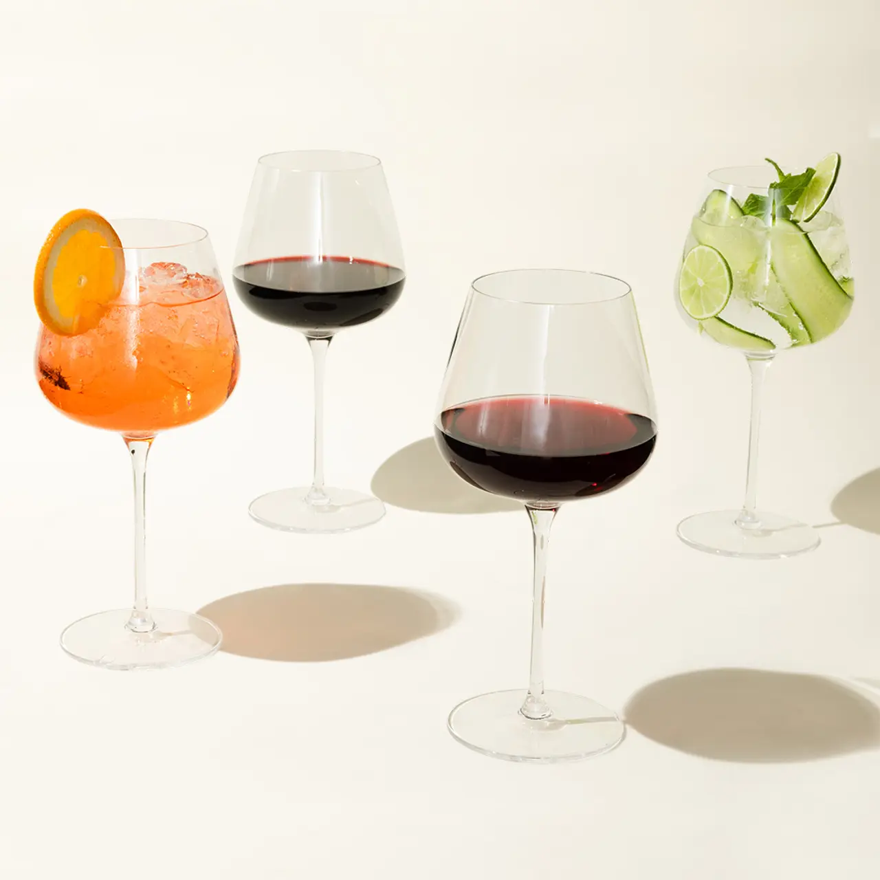 Four different types of drinks, including what appears to be a spritz, two red wines, and a gin and tonic, are displayed in elegant stemware against a neutral background.