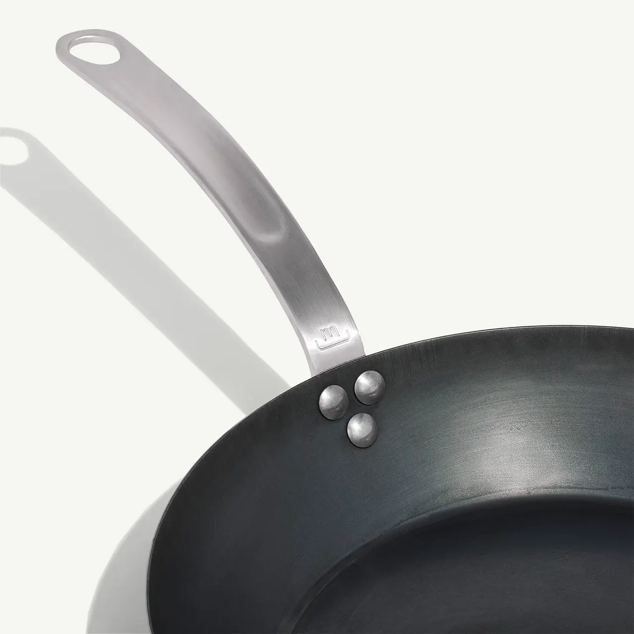 A close-up of two non-stick frying pans with stainless steel handles overlaps, casting a shadow on a light surface.